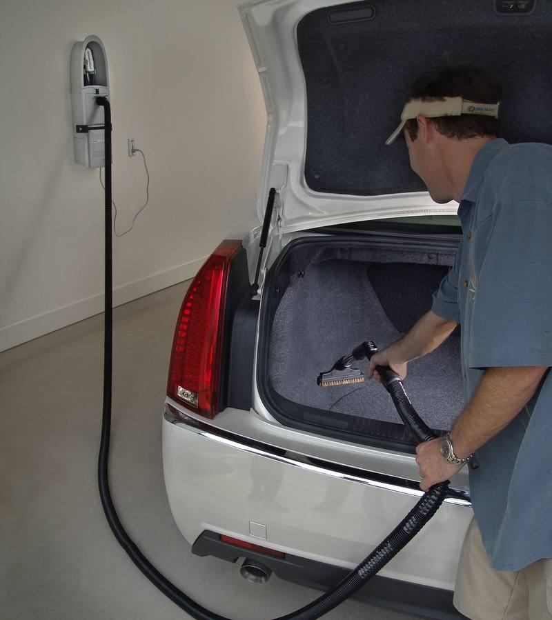 Someone vacuuming the carpeted trunk of a white sedan using the GarageVac hose run from the wall unit behind them.